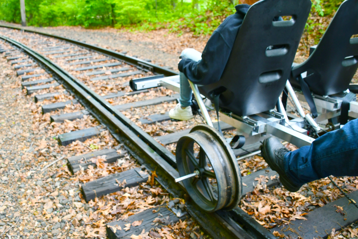 Two people on a pedal-powered rail vehicle on train tracks in a forest.