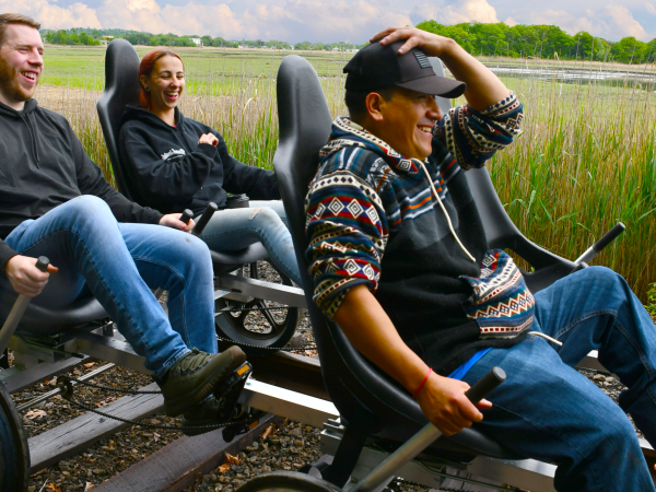 Three people laugh while riding rail bikes outdoors on a sunny day.